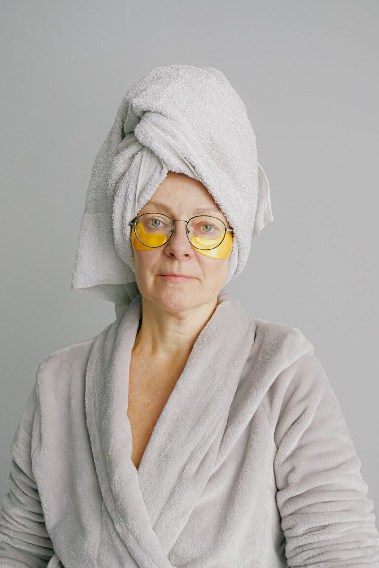 A mature woman over 50 wearing a bathrobe, a head towel, and gold under-eye patches while preparing her morning skin routine.